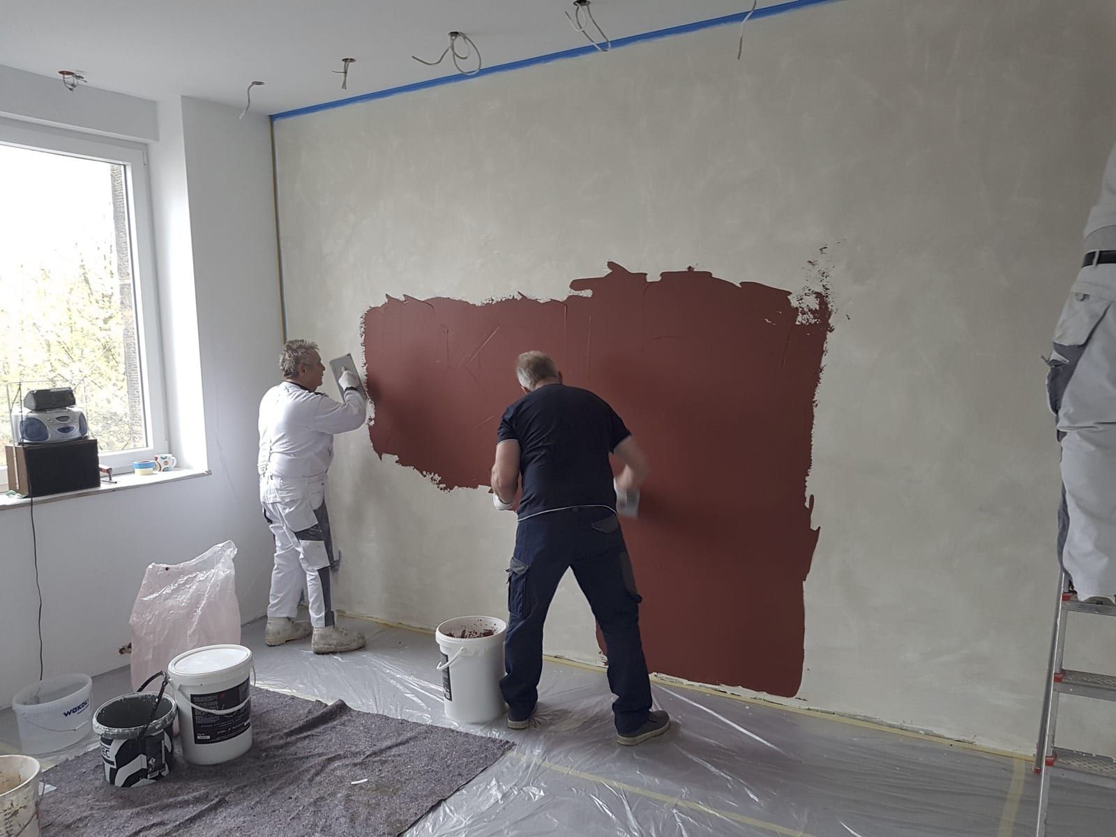 Zwei Mitarbeiter tragen roten Kalkedelputz auf die Wandfläche auf