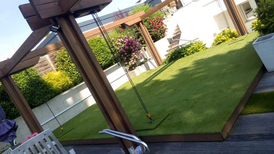 Terrasse mit Holzpergola und Holzdielenboden, daneben eine erhöhte Rasenfläche und blühende Sträucher